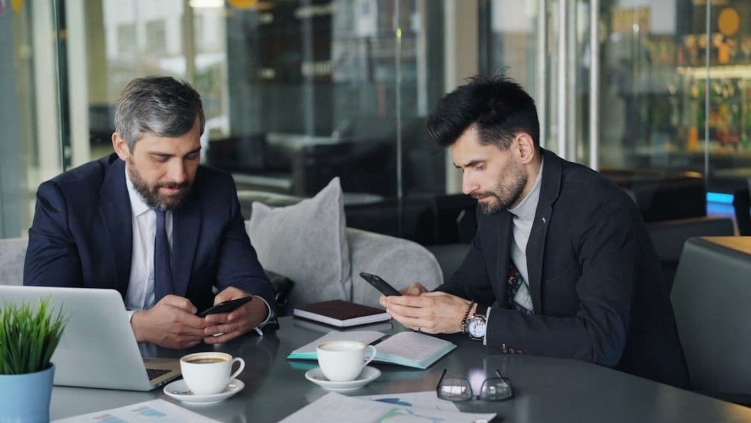 two men sitting at a table looking at their phones